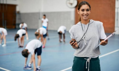 Young happy coach using stopwatch during PE class at school gym and looking at camera. Her students are exercising in the background.